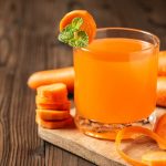 Vaso de jugo naranja brillante rodeado de zanahorias frescas, mitades de naranja y raíz de jengibre sobre una superficie blanca.