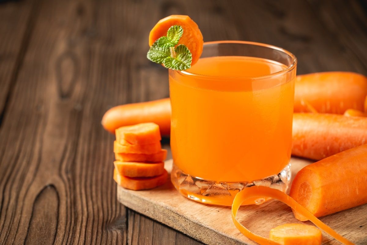 Vaso de jugo naranja brillante rodeado de zanahorias frescas, mitades de naranja y raíz de jengibre sobre una superficie blanca.