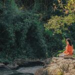Mujer sentada en posición de meditación sobre una roca junto a un río, rodeada de vegetación verde y luz natural.