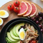 Plato keto con ensalada de aguacate, pollo desmenuzado y huevo cocido sobre una tabla de madera, junto a rodajas de toronja, tomates cherry y cerezas.