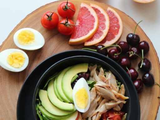 Plato keto con ensalada de aguacate, pollo desmenuzado y huevo cocido sobre una tabla de madera, junto a rodajas de toronja, tomates cherry y cerezas.
