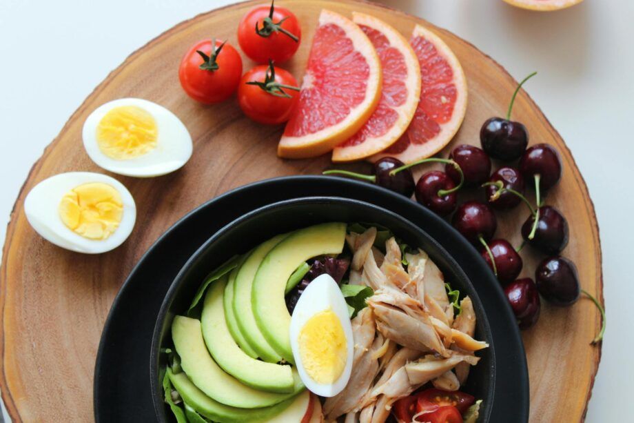Plato keto con ensalada de aguacate, pollo desmenuzado y huevo cocido sobre una tabla de madera, junto a rodajas de toronja, tomates cherry y cerezas.