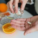 Manos vertiendo una cápsula de suplemento desde un frasco, con naranjas y blísters de vitaminas sobre la mesa, ilustrando el uso de citrato de magnesio.