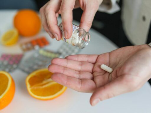 Manos vertiendo una cápsula de suplemento desde un frasco, con naranjas y blísters de vitaminas sobre la mesa, ilustrando el uso de citrato de magnesio.