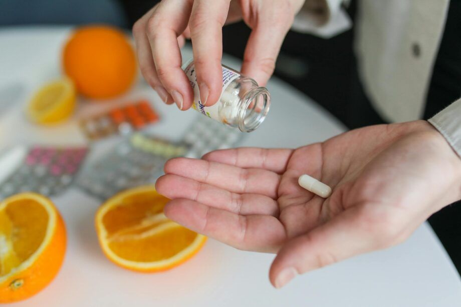 Manos vertiendo una cápsula de suplemento desde un frasco, con naranjas y blísters de vitaminas sobre la mesa, ilustrando el uso de citrato de magnesio.