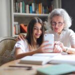 Abuela y nieta compartiendo un momento cercano mientras miran juntas un teléfono móvil en una mesa llena de libros. La abuela, con cabello canoso y gafas, muestra atención y cariño, mientras la nieta sonríe sosteniendo el dispositivo. La escena simboliza la conexión afectiva intergeneracional, la transmisión de sabiduría y valores familiares, y el rol de la abuela paterna en el apoyo emocional y la integración de la historia familiar, aspectos clave en las constelaciones familiares.