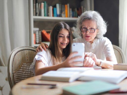 Abuela y nieta compartiendo un momento cercano mientras miran juntas un teléfono móvil en una mesa llena de libros. La abuela, con cabello canoso y gafas, muestra atención y cariño, mientras la nieta sonríe sosteniendo el dispositivo. La escena simboliza la conexión afectiva intergeneracional, la transmisión de sabiduría y valores familiares, y el rol de la abuela paterna en el apoyo emocional y la integración de la historia familiar, aspectos clave en las constelaciones familiares.