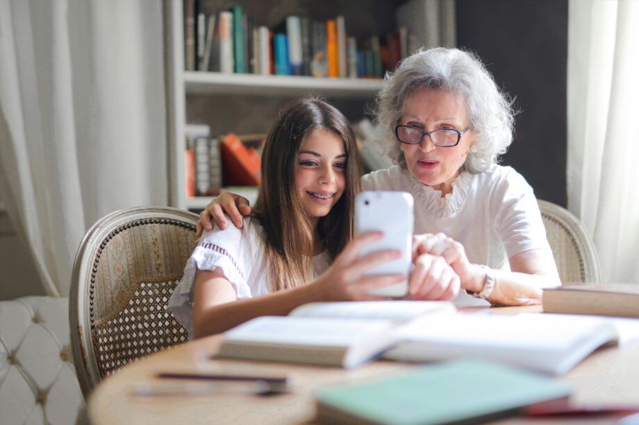 Abuela y nieta compartiendo un momento cercano mientras miran juntas un teléfono móvil en una mesa llena de libros. La abuela, con cabello canoso y gafas, muestra atención y cariño, mientras la nieta sonríe sosteniendo el dispositivo. La escena simboliza la conexión afectiva intergeneracional, la transmisión de sabiduría y valores familiares, y el rol de la abuela paterna en el apoyo emocional y la integración de la historia familiar, aspectos clave en las constelaciones familiares.
