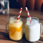 Dos batidos en frascos de vidrio con pajillas, uno de color mango y otro cremoso, que aparentan ser saludables pero pueden contener altos niveles de azúcar.