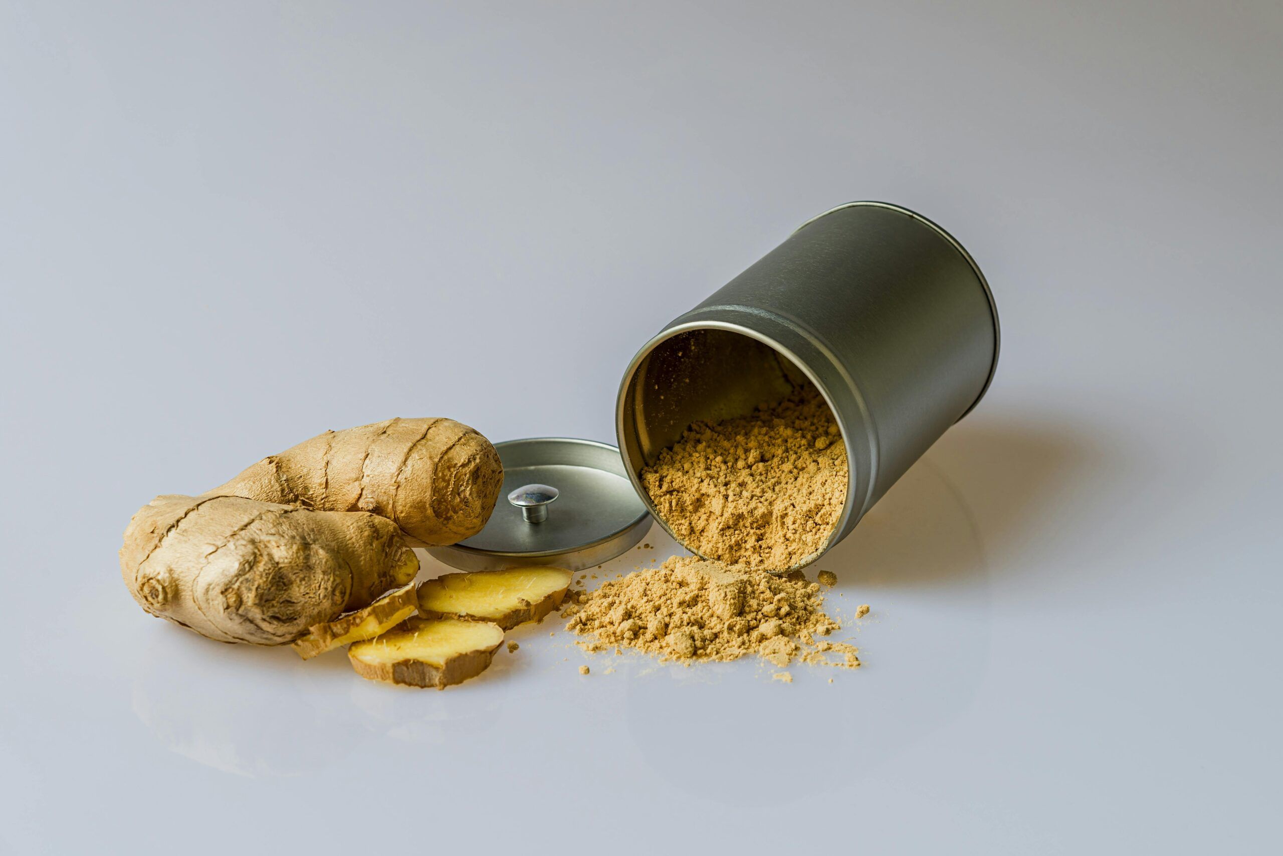 Raíz de jengibre fresco con rodajas y jengibre en polvo derramado de un recipiente, especia natural con propiedades antiinflamatorias.
