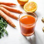Vaso de batido antioxidante de zanahoria, naranja y jengibre junto a sus ingredientes frescos sobre una mesa blanca.