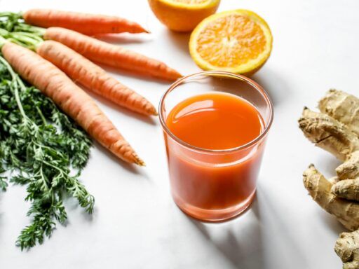 Vaso de batido antioxidante de zanahoria, naranja y jengibre junto a sus ingredientes frescos sobre una mesa blanca.