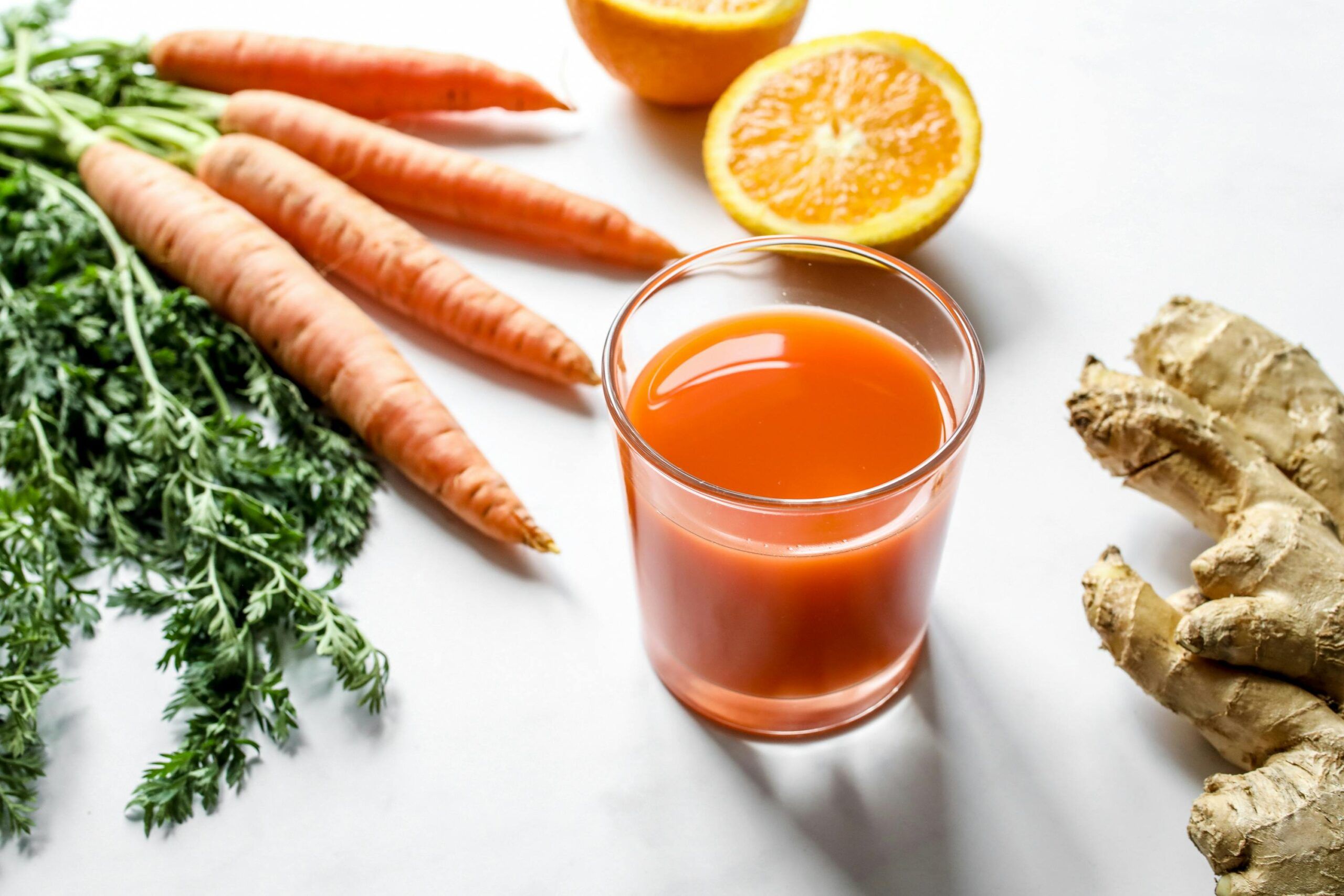 Vaso de batido antioxidante de zanahoria, naranja y jengibre junto a sus ingredientes frescos sobre una mesa blanca.