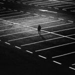 Figura solitaria de un hombre en un gran estacionamiento vacío iluminado en contraste, representando sentimientos de abandono y soledad emocional.