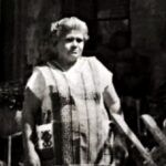 Mujer mayor con vestimenta tradicional de curandera, de pie en un espacio exterior con plantas y utensilios típicos del curanderismo.