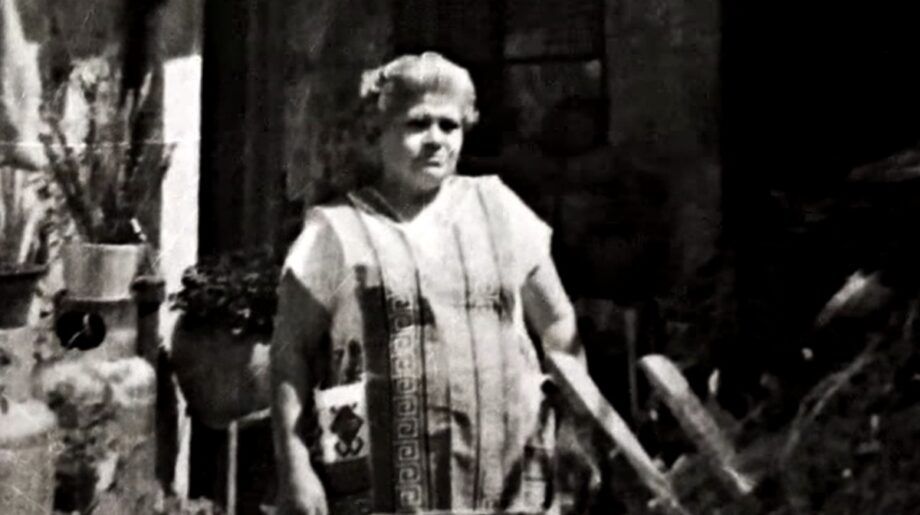 Mujer mayor con vestimenta tradicional de curandera, de pie en un espacio exterior con plantas y utensilios típicos del curanderismo.