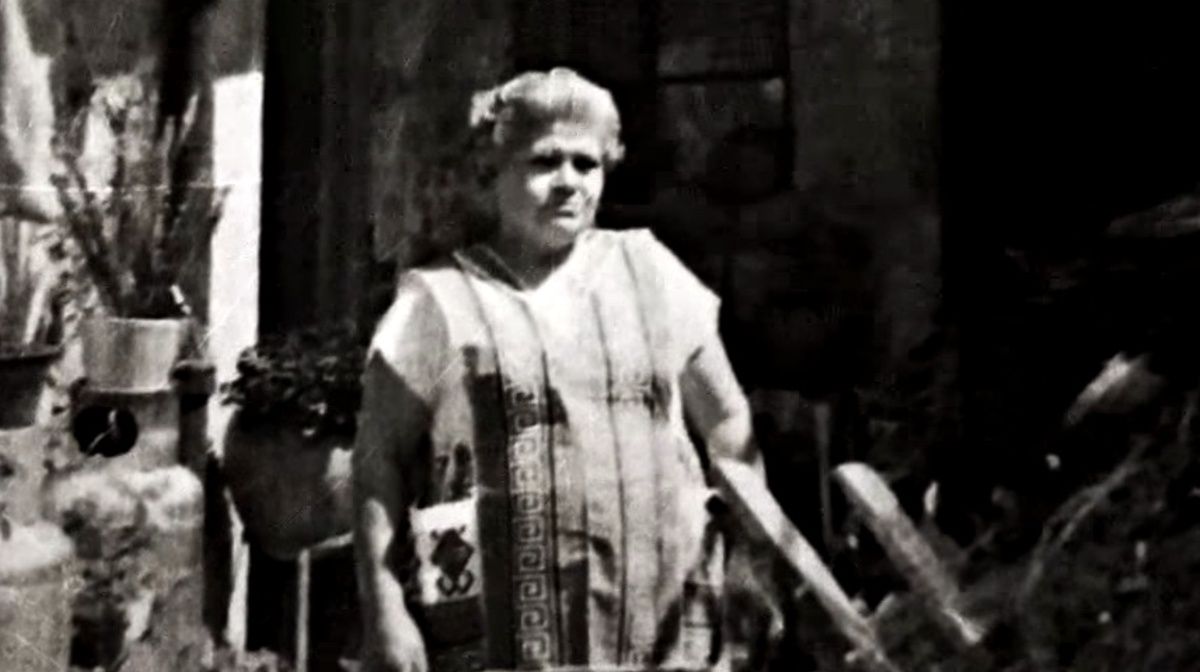Mujer mayor con vestimenta tradicional de curandera, de pie en un espacio exterior con plantas y utensilios típicos del curanderismo.