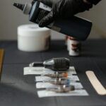Mano con guante negro sosteniendo un bote de tinta sobre una mesa con agujas, puntas esterilizadas y herramientas de tatuaje dispuestas ordenadamente.