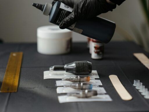Mano con guante negro sosteniendo un bote de tinta sobre una mesa con agujas, puntas esterilizadas y herramientas de tatuaje dispuestas ordenadamente.