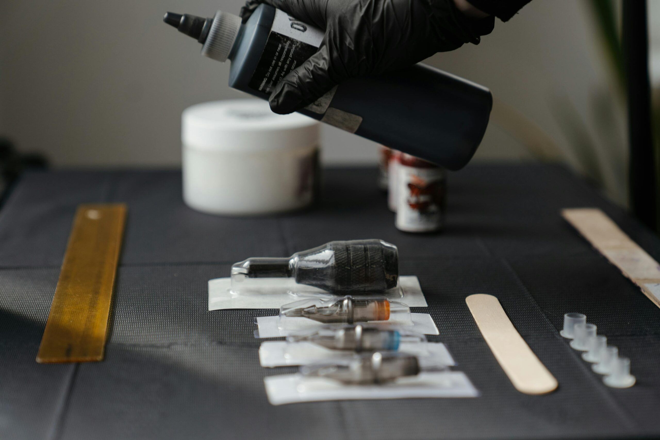 Mano con guante negro sosteniendo un bote de tinta sobre una mesa con agujas, puntas esterilizadas y herramientas de tatuaje dispuestas ordenadamente.