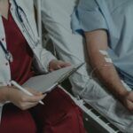 Profesional médico tomando notas junto a un paciente hospitalizado con una vía intravenosa en el brazo.
