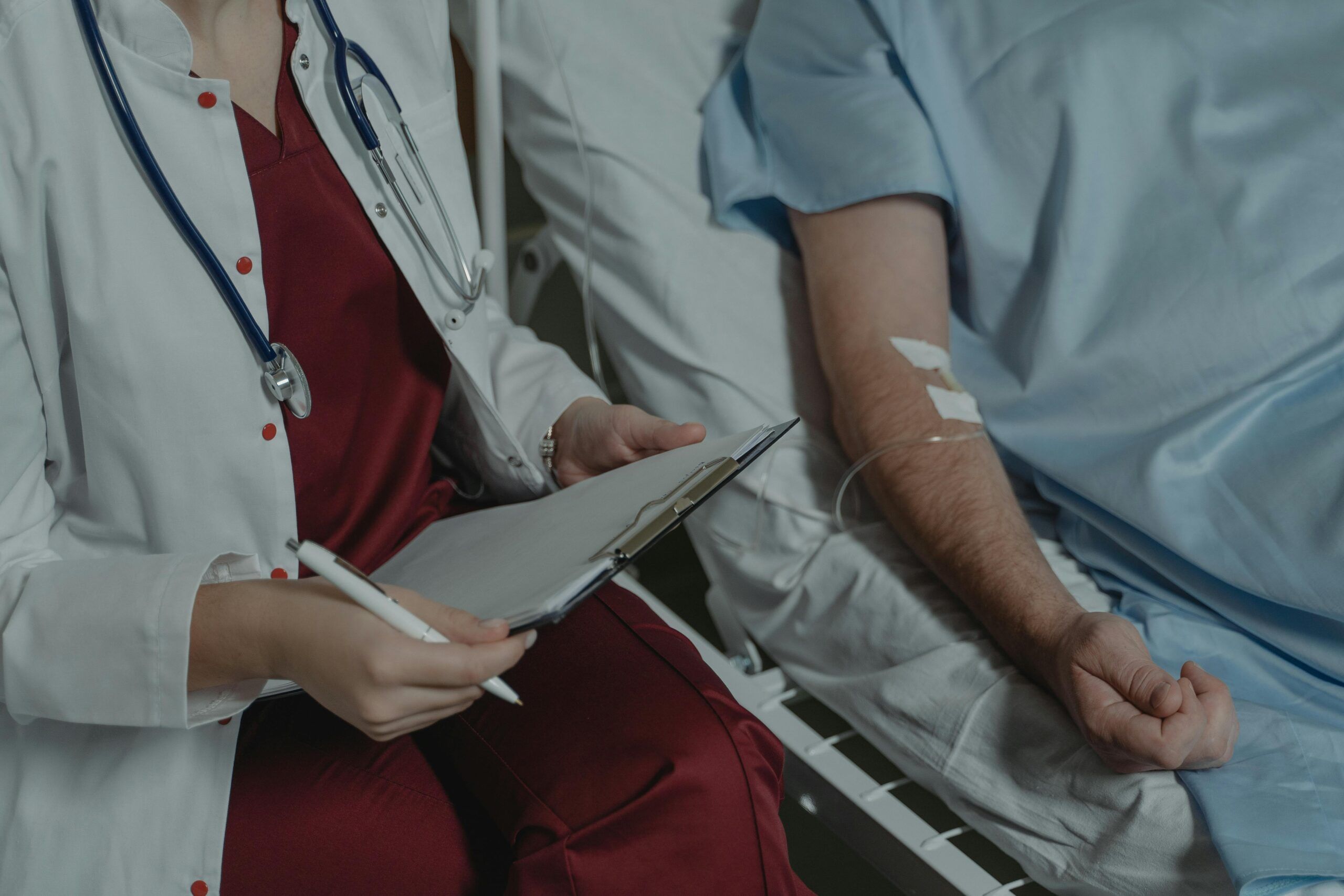 Profesional médico tomando notas junto a un paciente hospitalizado con una vía intravenosa en el brazo.
